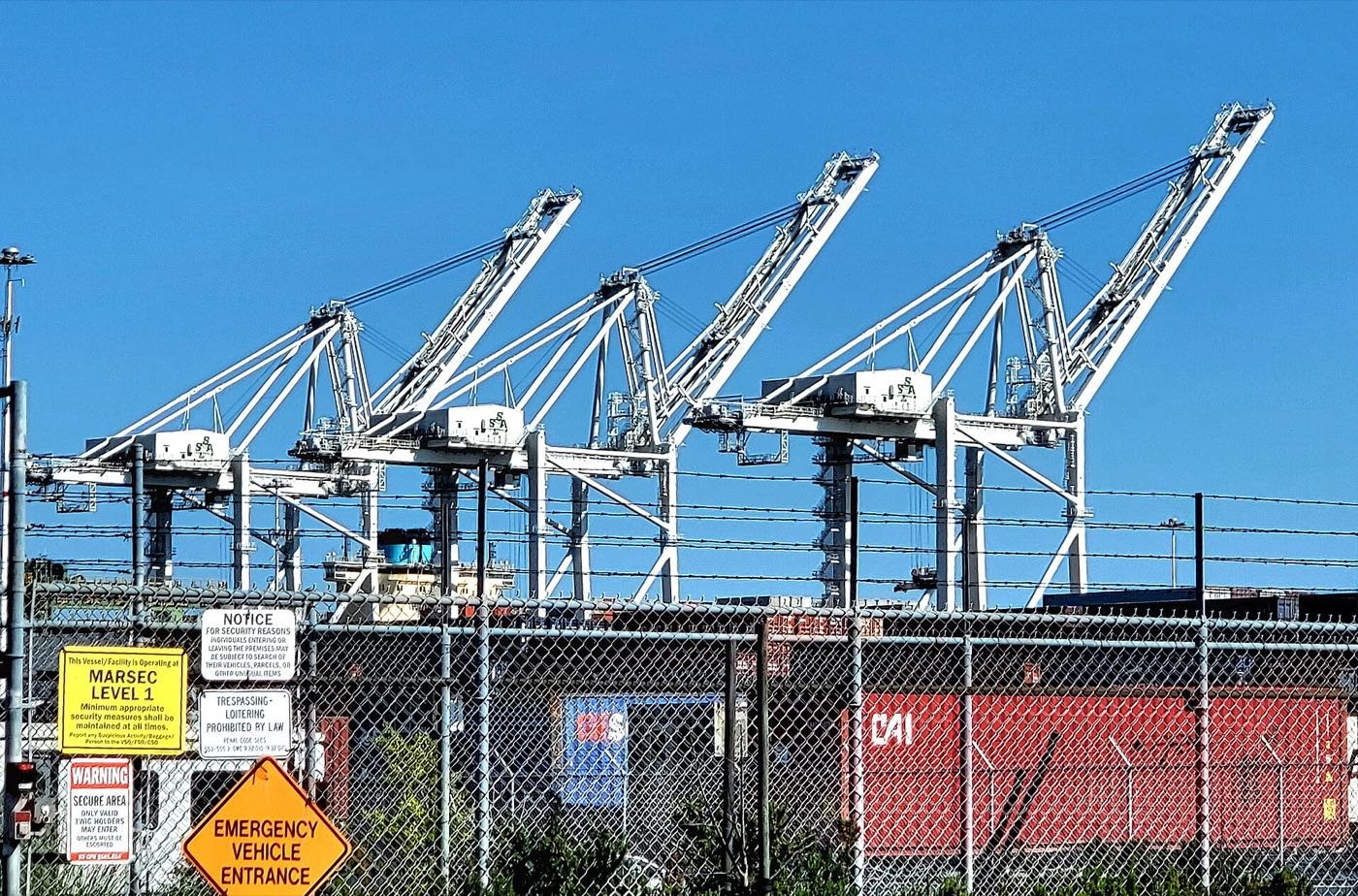 White container cranes against a blue sky above stacked shipping containers at the Port of Oakland